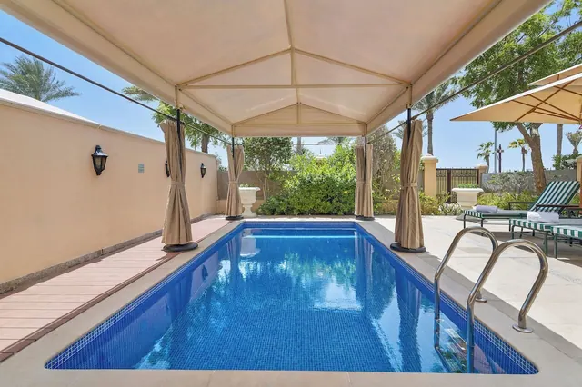 A private rectangular swimming pool sits under a cream canopy with hanging curtains, with striped sun loungers and an umbrella beside it.