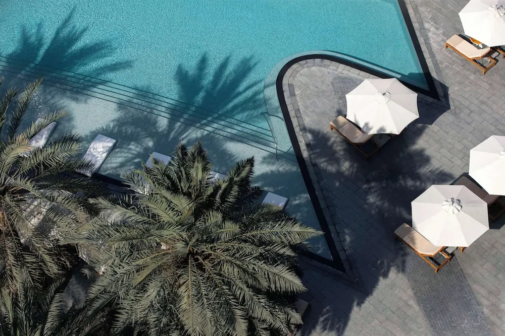 Aerial view of a turquoise swimming pool with white parasol umbrellas, sun loungers and palm trees casting shadows across the paving.