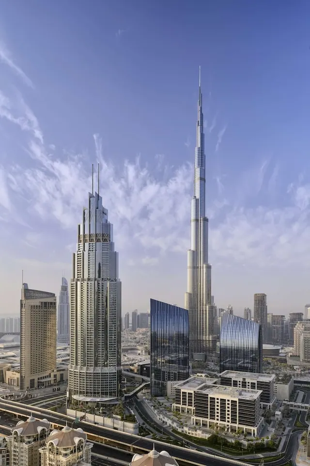 Elevated view of the Dubai skyline with the Burj Khalifa rising above surrounding towers and roads under a hazy blue sky.