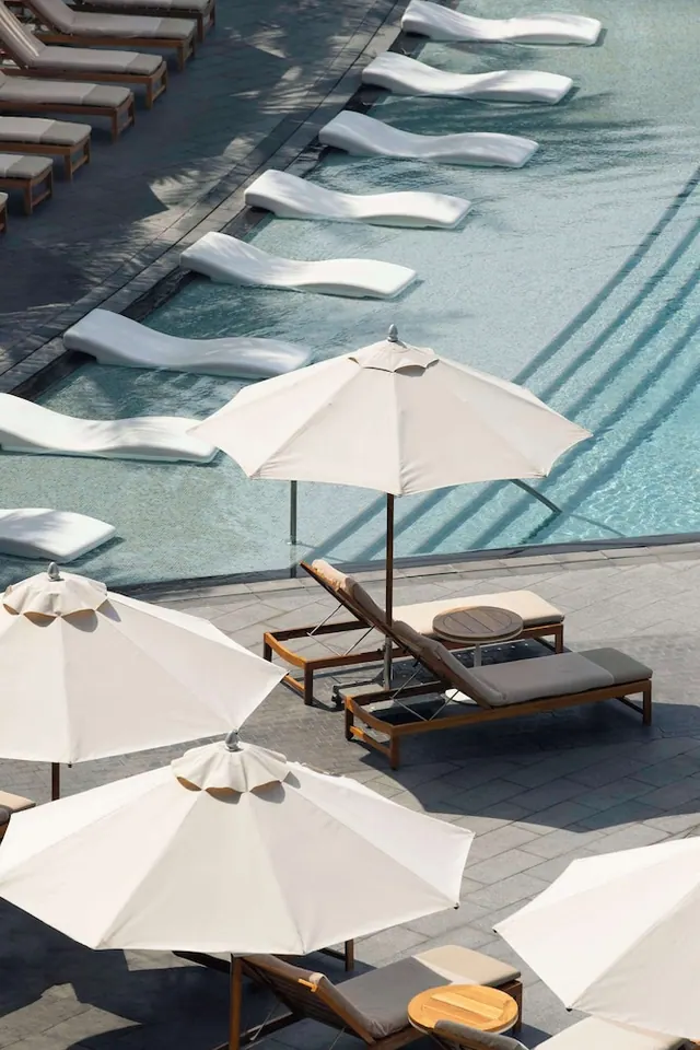 Elevated view of a pool with white floating loungers in the water, white parasols and wooden sun loungers with grey cushions on the paved deck.