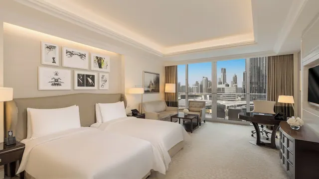 A twin-bedded hotel room with framed artwork, a seating area, a work desk and floor-to-ceiling windows overlooking a city skyline.