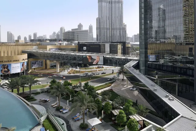 Elevated view over a pool with sun loungers and palm trees, a pedestrian skybridge and road traffic, with the Dubai Mall and the Burj Khalifa in the background.