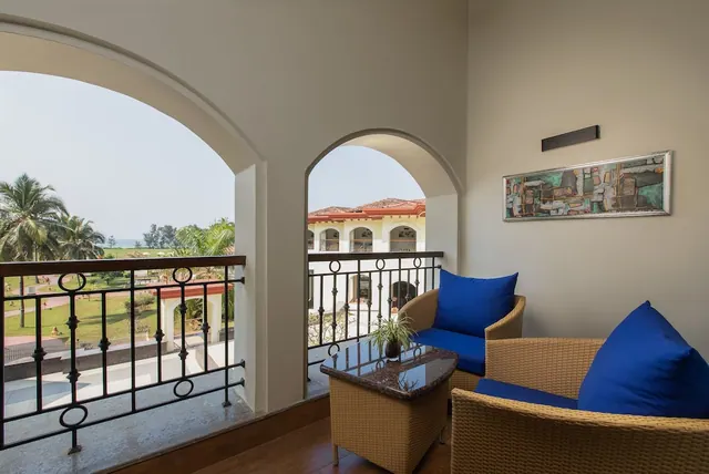 A covered balcony has wicker seating, blue cushions, and views of the landscaped grounds and sea beyond.