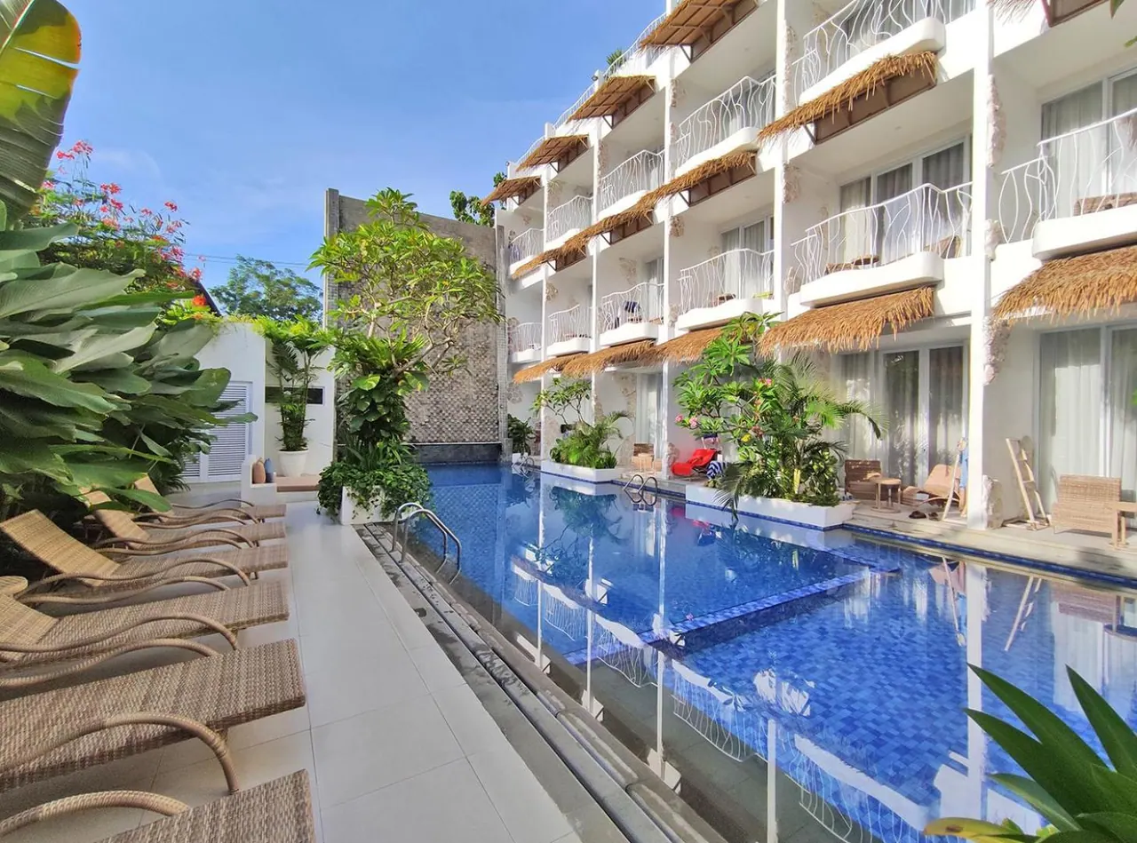 A rectangular blue-tiled swimming pool runs beside a white multi-storey building with thatched balcony awnings, and sun loungers line the pool deck on the left.