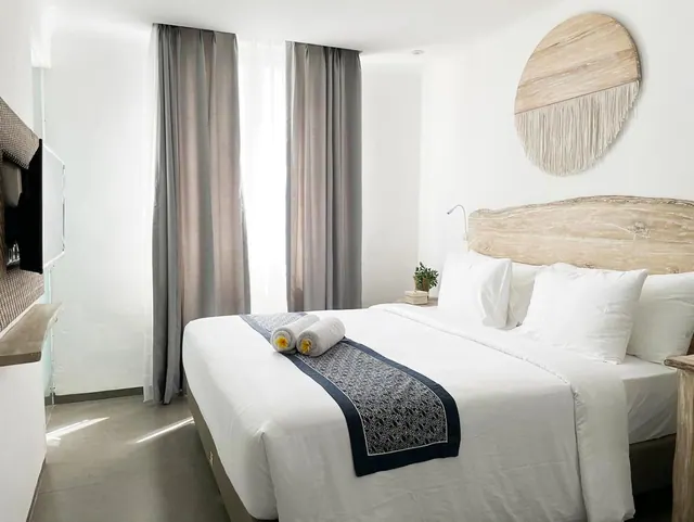 A bedroom with a made double bed with white linen, a patterned runner with two rolled towels, a wooden headboard, grey curtains at the window, and a wall-mounted television.