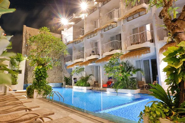 A night view of an illuminated swimming pool with wicker sun loungers in the foreground, tropical plants, and a multi-storey building with balconies under thatched roofs.