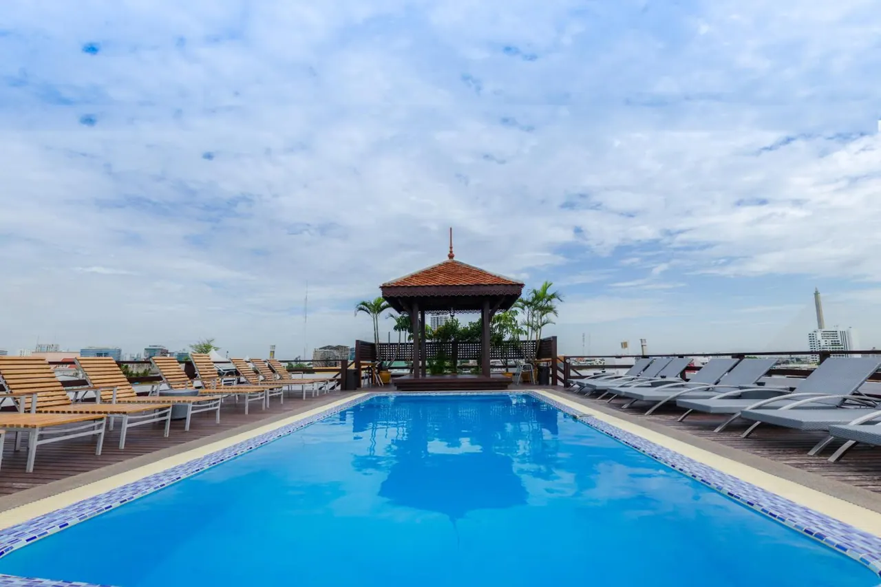 A rooftop swimming pool with blue water, wooden and grey sun loungers along its sides, and a Thai-style gazebo at the far end under a cloudy sky.