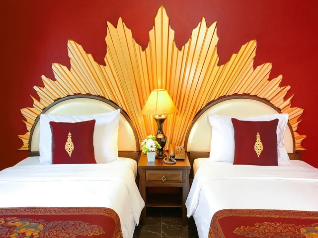 Two single beds with white linen and red cushions sit against a red wall with a gold fan-shaped headboard, with a bedside table, lamp and telephone between them.