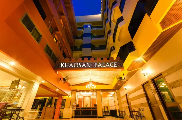Upward view of the Khaosan Palace building facade at dusk, with its illuminated sign and the lobby entrance below.