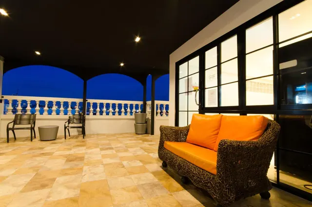 A covered terrace at dusk with an orange wicker sofa, metal benches, a tiled floor, and an arched balustrade.