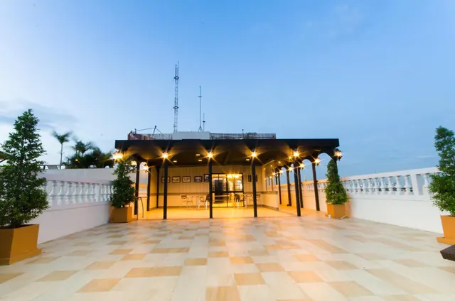 A covered rooftop terrace with an illuminated pavilion, tiled floor, white balustrade and potted trees at dusk.