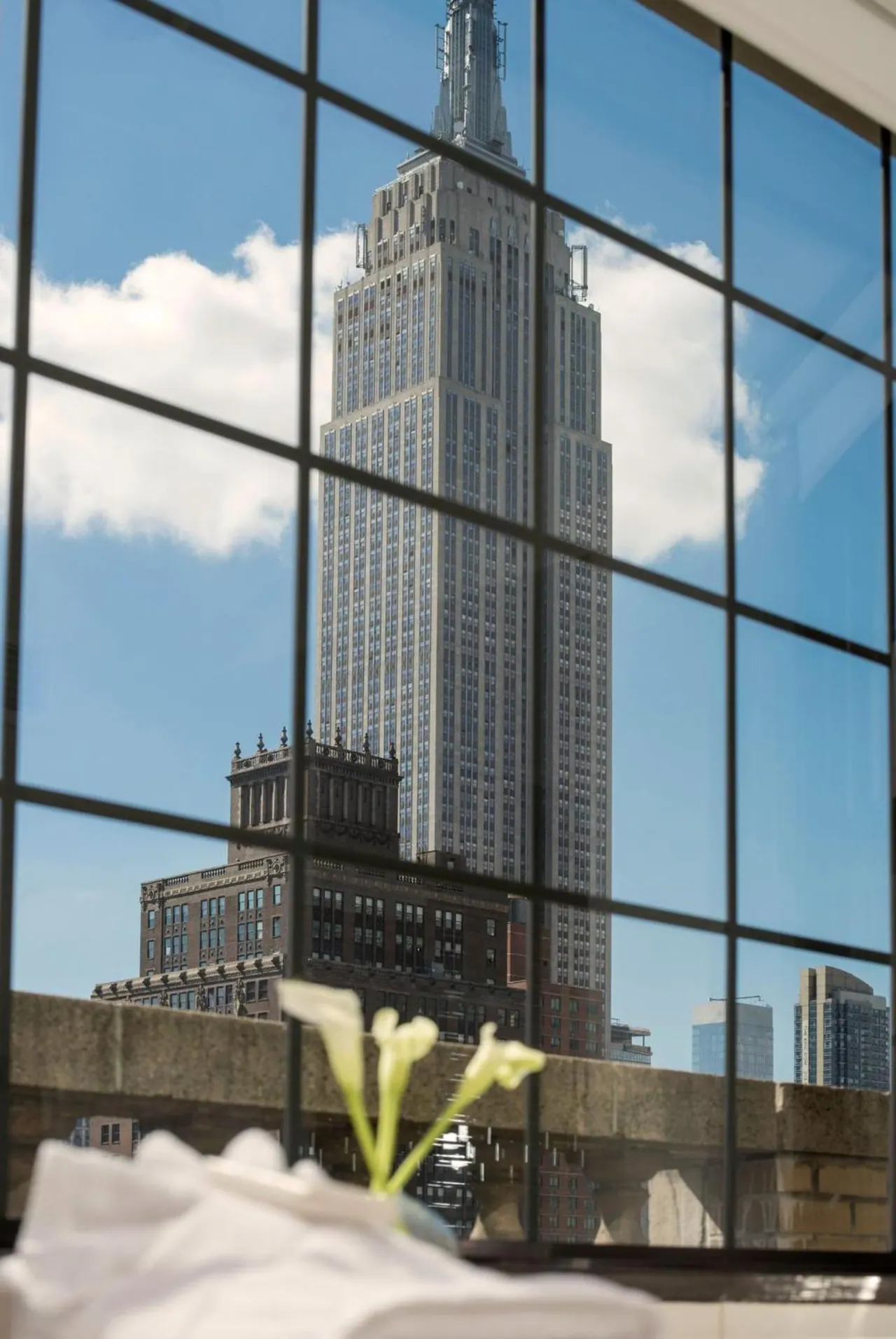 A view through a large grid-paned window of the Empire State Building and nearby buildings, with white flowers and linen in the foreground.