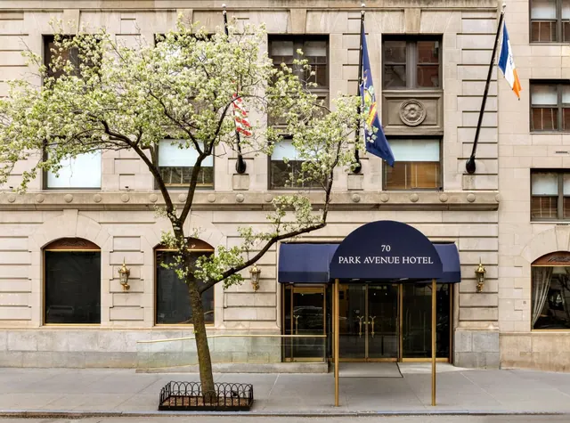 The limestone front of the Park Avenue Hotel with a navy awning, glass entrance doors and a flowering tree on the sidewalk.