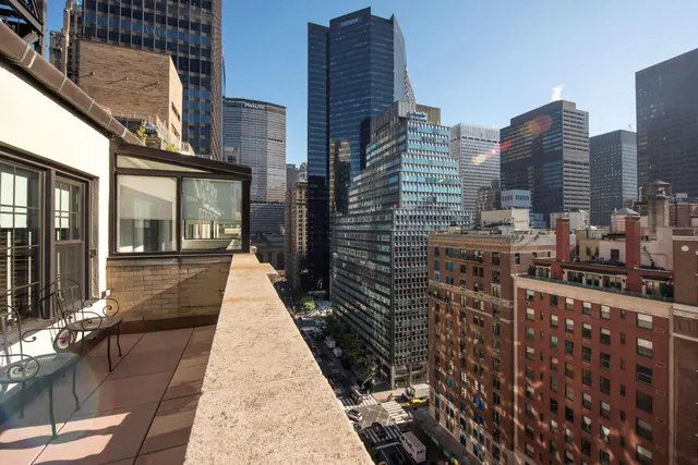 A rooftop terrace with metal chairs and a small table overlooks dense midtown skyscrapers and street traffic below.