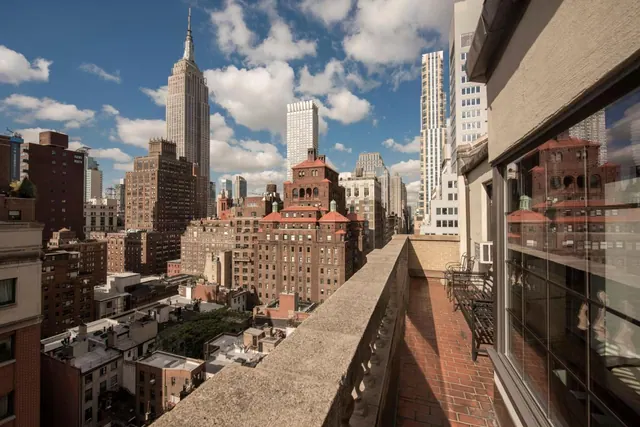 A brick-floored terrace with a stone railing and metal chairs overlooks midtown Manhattan, with the Empire State Building visible in the distance.