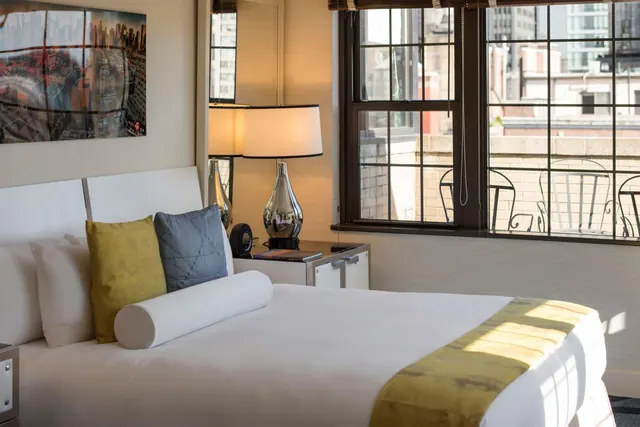 A bedroom with a made white bed, bedside lamps, and a large window looking out onto city buildings and a small balcony with metal chairs.