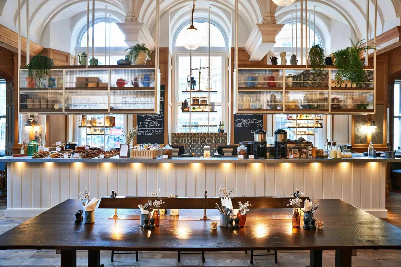 A café interior with a long wooden communal table in front of a counter holding pastries and coffee equipment, under arched ceilings and tall windows.