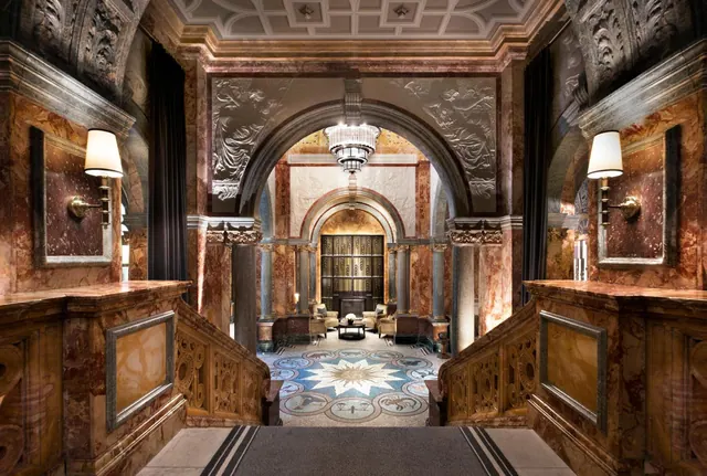 A marble-lined lobby with a grand staircase, a patterned mosaic floor, and an arched doorway leading to a seating area beneath a chandelier.