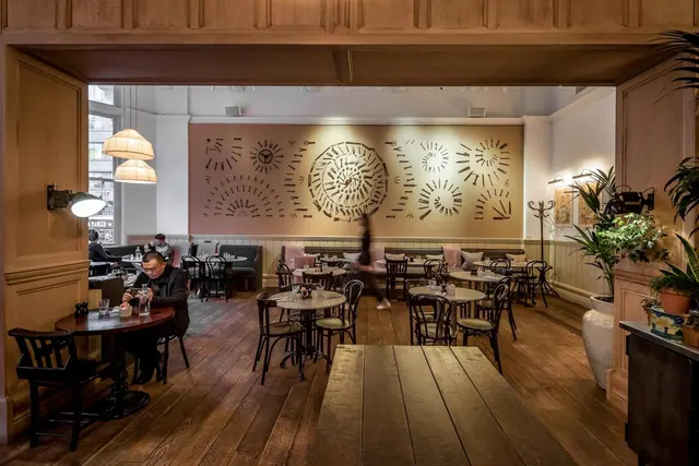 A dining room with wooden panelling, round wooden tables and bentwood chairs, a large patterned mural on the back wall, and a few people seated at tables.