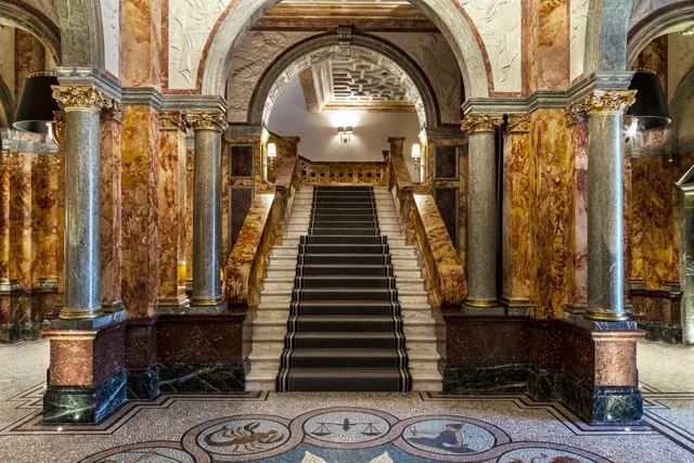 A grand marble staircase with a dark runner leads up under a decorated arch in a lobby with a mosaic floor and marble columns.