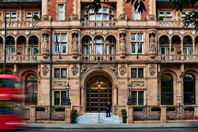 The ornate stone and brick facade of a hotel with a central arched entrance, steps, and a uniformed doorman, with a blurred red bus passing in the foreground.