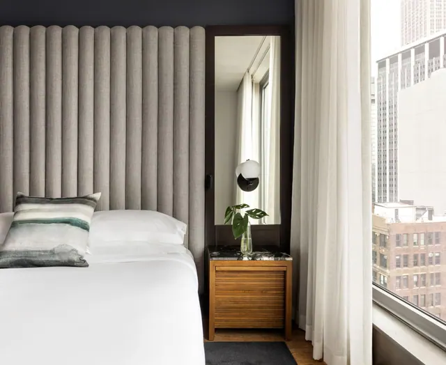A bedroom with a tufted grey upholstered headboard, a made bed with white linen, a marble-topped wooden nightstand with a lamp and plant, and a window overlooking city buildings.