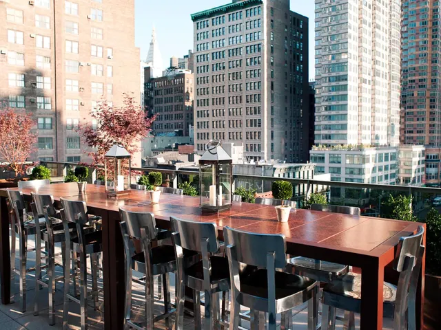 A rooftop terrace with a long wooden table, metal bar stools, lanterns and potted plants, overlooking high-rise buildings.