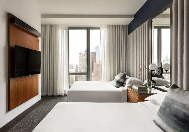 A hotel bedroom with two made beds, a wall-mounted television, and a window looking out over city buildings.