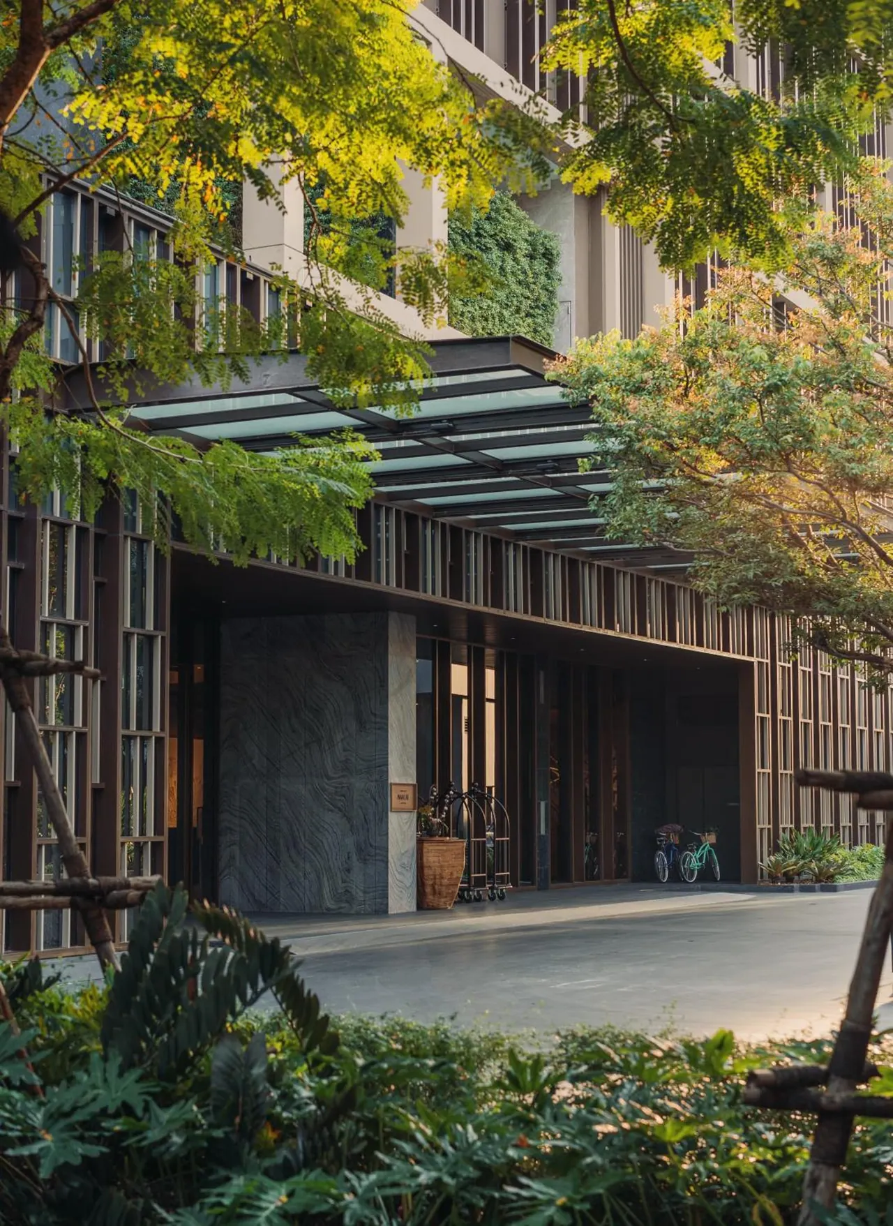 The covered entrance of a hotel building with a glass canopy, a luggage trolley, a large planter and two parked bicycles, framed by trees.