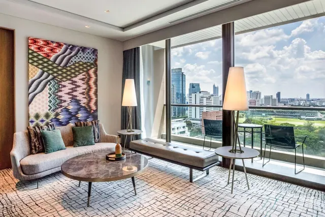 A suite living area with a grey sofa, marble coffee table, padded bench and floor lamps, with floor-to-ceiling windows opening onto a balcony and a city skyline.