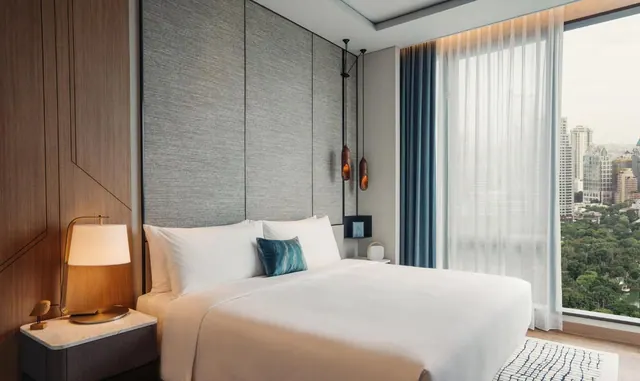 A bedroom with a made king bed, a bedside lamp and pendant lights, and a floor-to-ceiling window looking over treetops toward city towers.