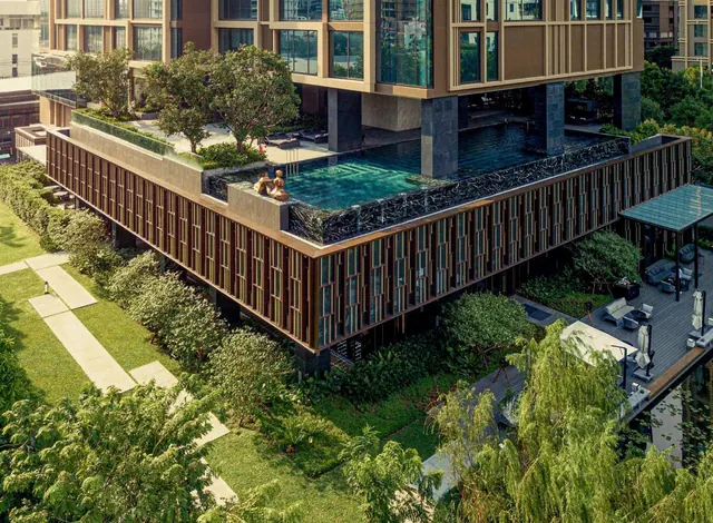 Aerial view of a hotel building with an infinity pool on a raised deck, surrounded by trees, a lawn with a stone path, and a covered lounge terrace.
