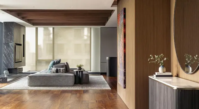 A living room with a grey sectional sofa, a rug on a wooden floor, slatted wood walls, a round mirror and a console table, with a large window covered by sheer blinds.