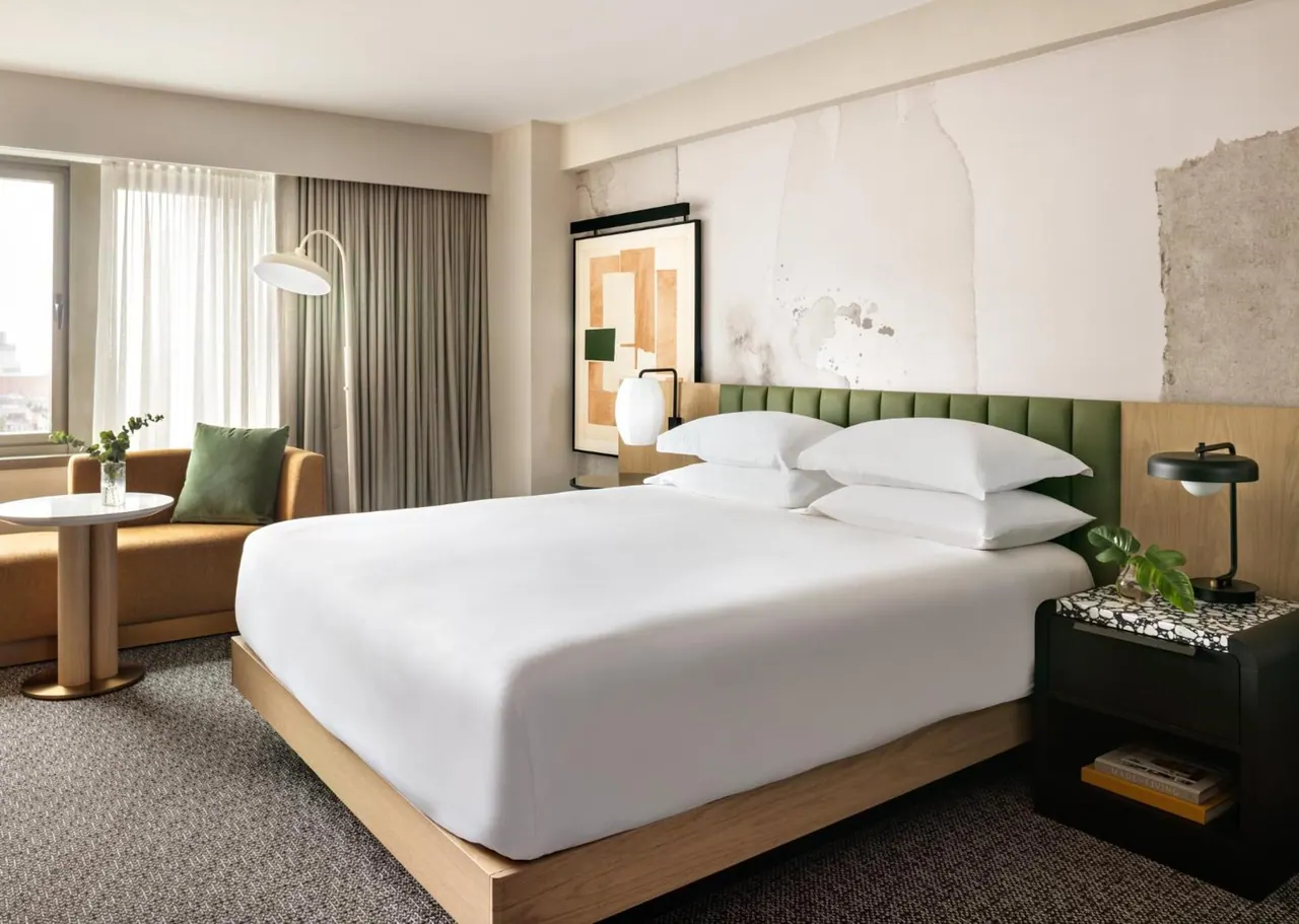 A king bed with white linens and a green tufted headboard sits in a hotel room beside an orange sofa, a round side table and an arc floor lamp.