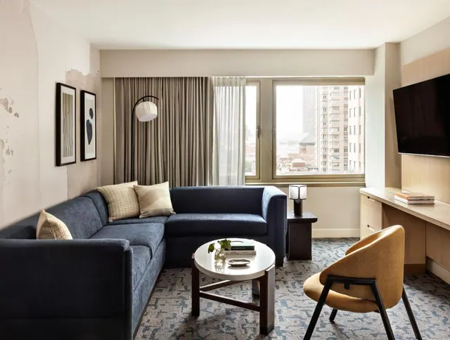 A living area with a navy sectional sofa, a round coffee table, an arc floor lamp, a wall-mounted television, and a window looking onto city high-rises.