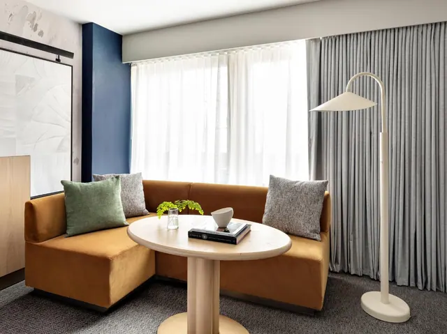 A seating area with an L-shaped mustard velvet banquette, a round light wood table holding a book and a small plant, a cream floor lamp, and grey curtains beside a window.