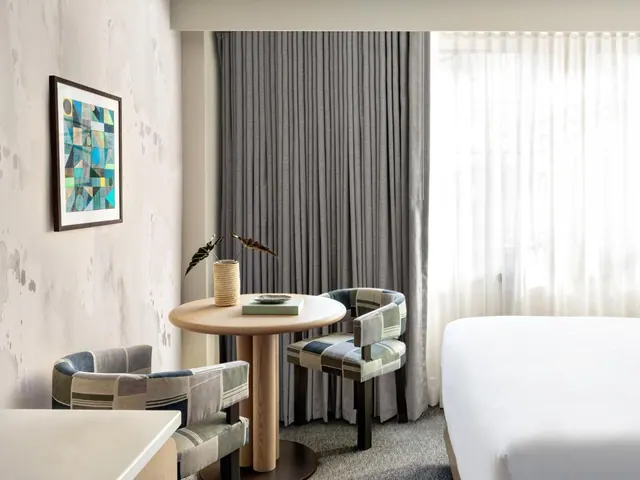 A hotel bedroom with a round wooden table, two upholstered chairs, grey drapes over a window with sheer curtains, and a made white bed at right.