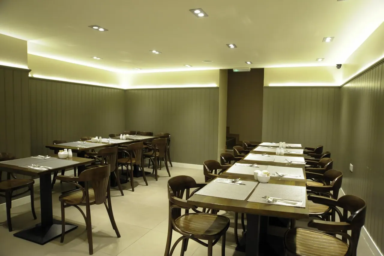 A restaurant dining room with wooden chairs and tables set with placemats, cutlery and condiment pots under ceiling downlights.