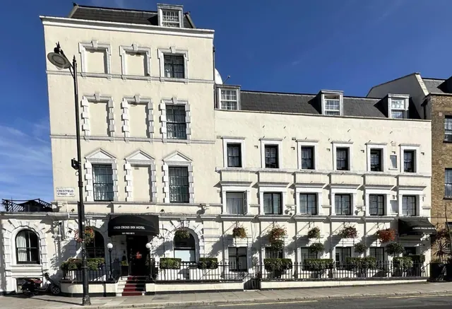 The cream-coloured front of the Kings Cross Inn Hotel faces the street under a clear blue sky, with a black awning, hanging flower baskets, a black railing and a street lamp.