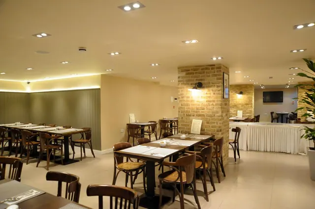An empty dining room with wooden bentwood chairs and set tables, a brick pillar with wall lights, a potted plant on the right, and a wall-mounted television in the background.