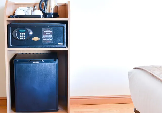 A wooden bedside unit holds a kettle, a tray with cups, a digital in-room safe and a small fridge, with the edge of a bed visible at right.