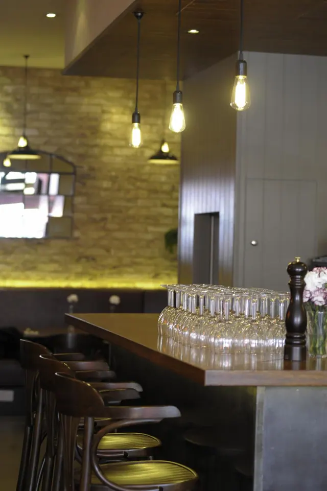 A bar counter holds rows of upturned wine glasses, a pepper mill and a vase of flowers, with bar stools in front and exposed-bulb pendant lights above a brick wall.