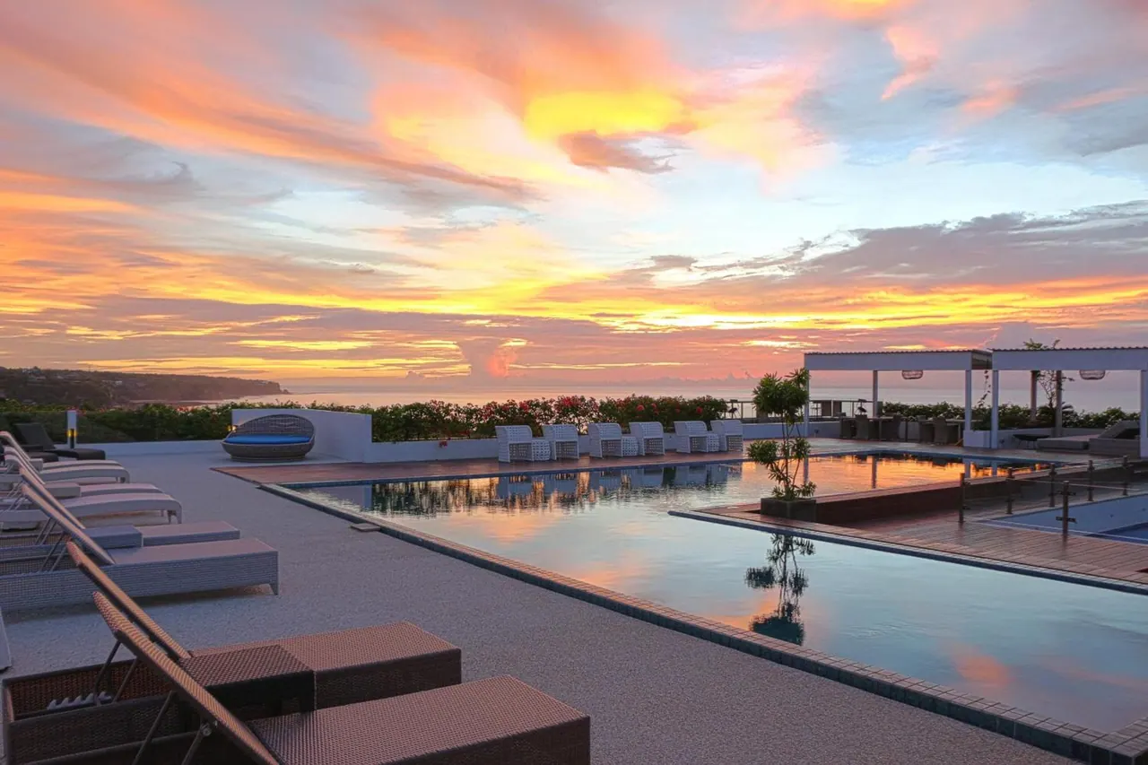 A long rectangular pool with white sun loungers on an outdoor deck overlooks the sea under a sunset sky.