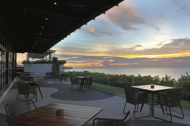 An outdoor dining terrace with grey chairs and wooden tables overlooks the ocean at sunset.