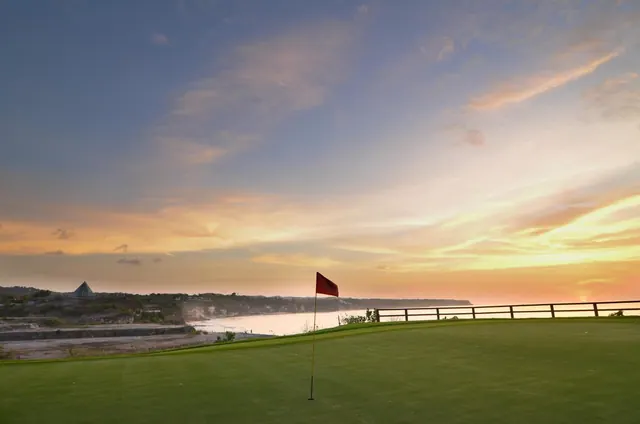 A golf green with a red flag sits beside a wooden rail on a cliff-top course overlooking the sea at sunset.