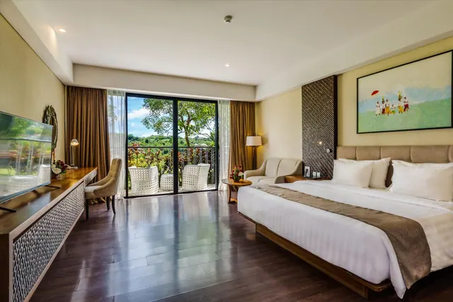 A bedroom with a king bed, an armchair, a wall-mounted television and a glass sliding door opening onto a balcony with white wicker chairs and a view of trees.