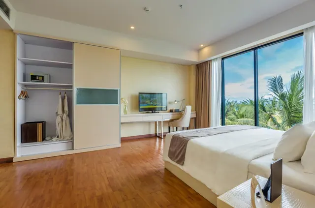 A bedroom with a large white bed, a wardrobe with a safe, a desk with a television, and a floor-to-ceiling window looking out onto palm trees.