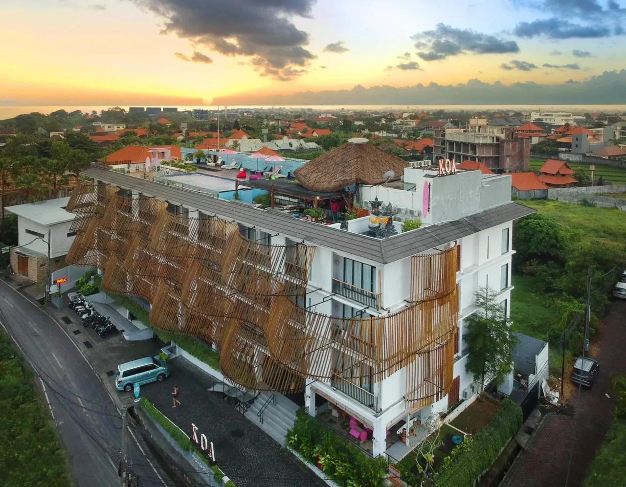 Aerial view at sunset of a white multi-storey hotel building with bamboo screens on its facade, a pool area with loungers and umbrellas, and the KOA sign on the roof, surrounded by neighbouring buildings.