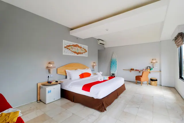 A bedroom with a king bed dressed in white and red linens, a wall map of Bali, a surfboard, a work desk with a kettle, and a wall-mounted air conditioner.