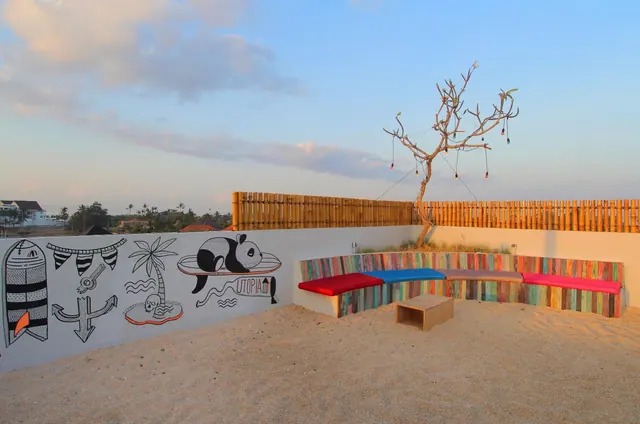 A sandy outdoor area with a white wall painted with surfboard, anchor, palm tree and panda drawings, a colourful wooden bench with red and blue cushions, and a bare tree beside a bamboo fence.